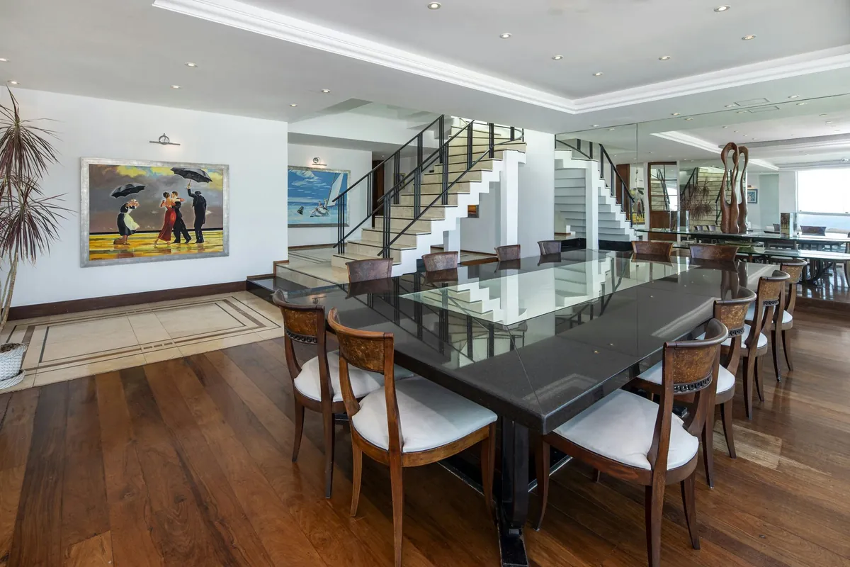 Long dining table and a staircase in the Copacabana duplex penthouse, with art on the walls and windows to the sea