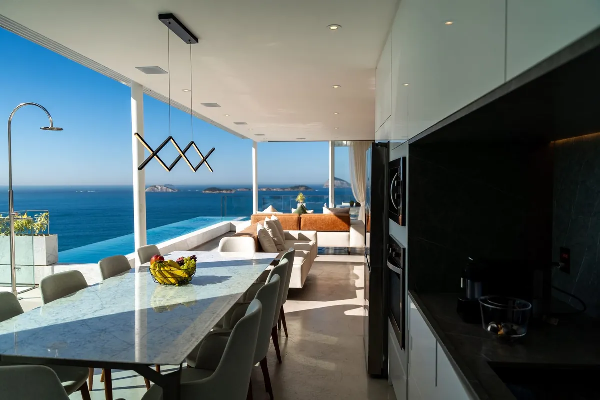 Dining table and kitchen of the Vidigal duplex penthouse with glass walls over the pool and the ocean