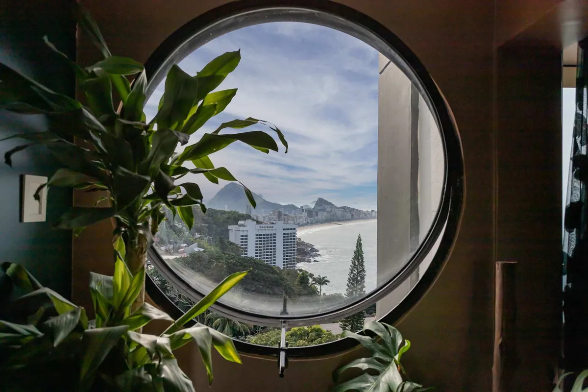 Round window in the Vidigal Sea-View Loft framing the Sheraton, Leblon and the coast, with a large plant beside it